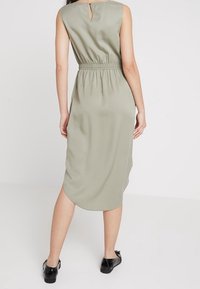 Sukienka bez rękawów w jasnym oliwkowym kolorze, z wycięciem na plecach, elastyczną talią i zaokrąglonym dołem. Materiał wydaje się gładki i lekko opływowy.