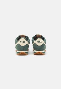 Zelené semišové tenisky s béžovým podpatkem, s logem "NIKE" na zadní straně. Gumová podešev s texturovaným vzorem pro lepší grip.