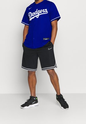 Blauw Los Angeles Dodgers shirt met witte letters, gecombineerd met zwarte shorts met witte strepen, en zwarte sneakers met groene accenten.