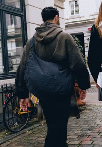 Černá nylonová taška přes rameno s texturovaným povrchem, nošena přes rameno. Muž v hnědé mikině jde po dlažební ulici, v pozadí kolo.