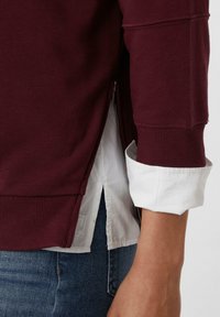 Bordeaux sweatshirt met geribbelde manchetten en zoom, met een zijritsdetail over een wit shirt daaronder, gecombineerd met blauwe jeans.