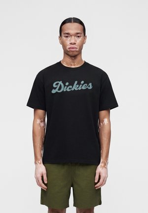 Άνδρας με λεύκη που φορά μαύρο t-shirt "Dickies" και χακί κοντό παντελόνι, στέκεται μπροστά σε μια απλή ανοιχτογκρι φόντο.