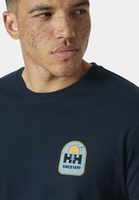 Camiseta azul marino de algodón, con un logo circular que presenta un árbol y un sol en amarillo y azul, con el texto "DESDE 1877" debajo.