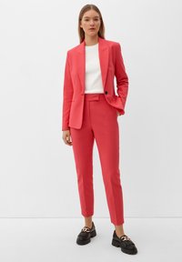 Blazer rose et pantalon sur mesure assorti, tous deux avec une texture lisse. Associés à un haut blanc et des chaussures noires avec des éléments décoratifs.