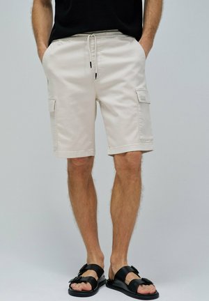 Pantalones cortos de carga beige con cintura elástica, bolsillos laterales y dobladillos cosidos; combinados con sandalias negras con correas ajustables.