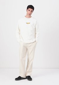 Hvit langermet skjorte med en liten gul grafikk, kombinert med beige bukser og svarte sko. Stoffet ser mykt og glatt ut.
