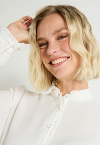 Blusa blanca con cuello con volantes y tejido texturizado. Presenta botones frontales con detalles circulares blancos. La modelo sonríe, mostrando la camiseta.