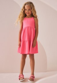 Różowa sukienka bez rękawów z teksturowanej tkaniny, z warstwowym krojem i rozkloszowaną spódnicą. Do niej dopasowano różowe sandały z kokardkami.
