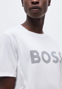 Κοντινό πλάνο ενός ατόμου που φοράει λευκό T-shirt με την επιγραφή «BOSS» σε μαύρες οριζόντιες ρίγες στο στήθος.