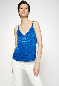 Top azul sem mangas com decote em V e um padrão de riscas de zebra texturizado. Feito de tecido leve, combinado com calças brancas.