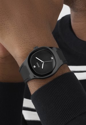 Black wristwatch with textured face and silver hands on wrist, worn over a dark ribbed sweater sleeve with white and black stripes.