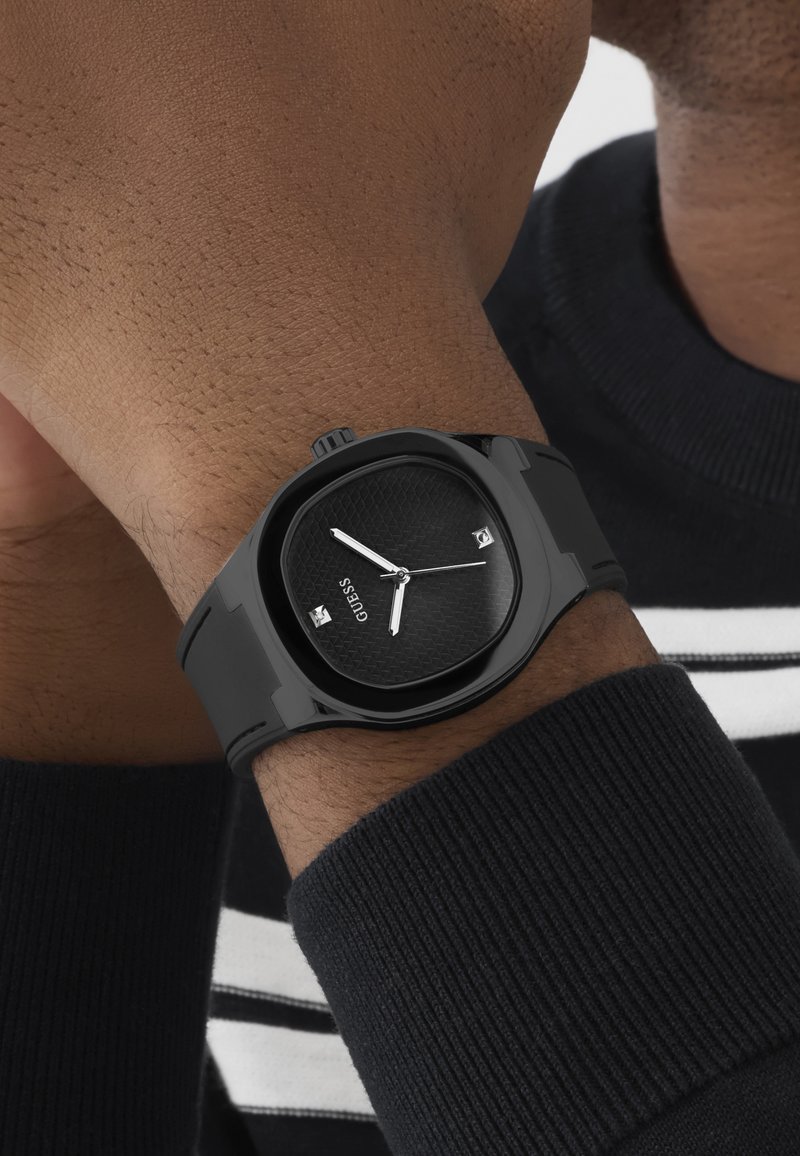 Montre-bracelet noire avec cadran texturé et aiguilles argentées portée au poignet, sur une manche de pull en côte sombre avec des rayures blanches et noires.