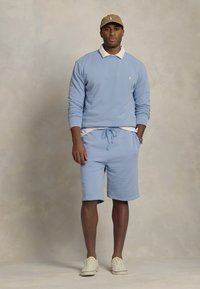 Sweatshirt azul claro com gola redonda, calções a condizer e uma camisola branca por dentro, combinados com um boné bege e sapatilhas brancas. Design simples.