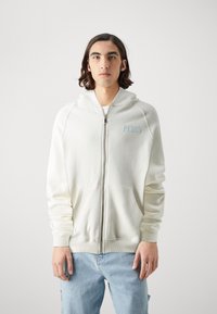Felpa bianca con zip e maniche raglan, texture morbida, tasca frontale e scritta azzurra "PEOUS" sul lato sinistro del petto. Indossata con jeans azzurri chiari.