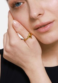 Goldring in Herzform mit glatter Oberfläche. Wird an einem Finger getragen, während eine Hand sanft das Gesicht berührt, was das Design hervorhebt.