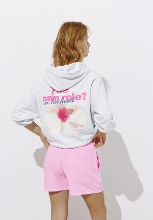 Kvinde med bølget blondt hår iført en hvid hættetrøje med blomstermotiv og tekst, kombineret med lyserøde shorts, stående foran en ensfarvet hvid væg.