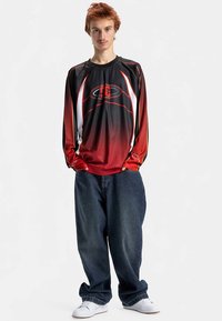 Hosszú ujjú mez fekete és piros árnyalatú dizájnnal, fehér részletekkel és logóval. Bő szabású sötét farmerrel és fehér cipővel párosítva.