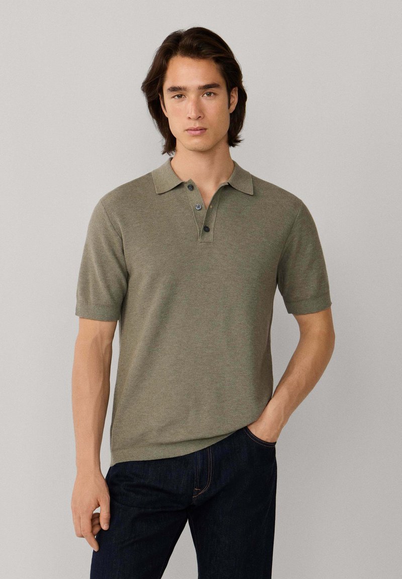 Jeune homme aux cheveux bruns portant un polo à manches courtes vert olive et un jean bleu foncé, debout avec une main dans la poche.