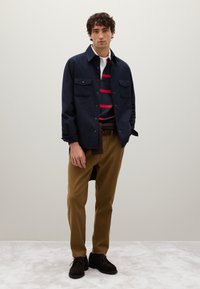 Hombre de pie que lleva una camisa azul marino, un suéter a rayas azul marino y rojo sobre una camisa blanca con cuello, pantalones marrones y zapatos marrones oscuros sobre un suelo gris.
