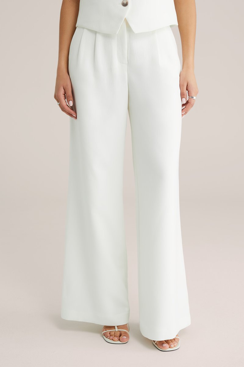Femme portant un pantalon blanc taille haute et jambes larges, associé à des sandales blanches à brides, debout devant un fond uni.