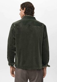 Hombre con cabello rizado, vistiendo una chaqueta de pana verde oscuro y pantalones grises, de espaldas contra un fondo claro liso.