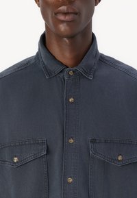 Temno denim srajca z ovratnikom, dvema žepoma na prsih in rjavimi gumbi. Ima teksturirano blago z rahlo izbledelim zaključkom.