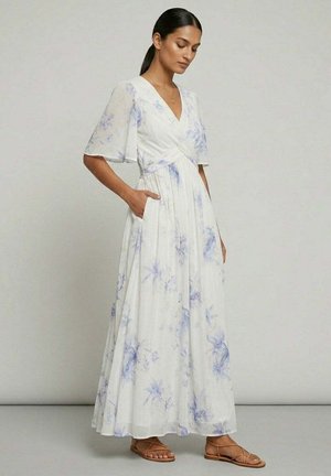 Femme portant une robe maxi blanche avec des motifs floraux bleus, col en V, manches volantées, poches latérales, et sandales plates marron, debout de profil.