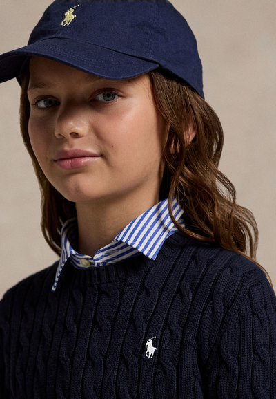 Noor inimene, kes kannab tumesinist mütsi ja kaabelmustrit kampsunit valge polo logoga, selle all on sinise ja valge triibulise kraega särk.