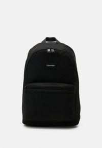 Zaino in tessuto nero con parte superiore arrotondata, doppie cerniere, tasca frontale, logo Calvin Klein e spallacci regolabili.