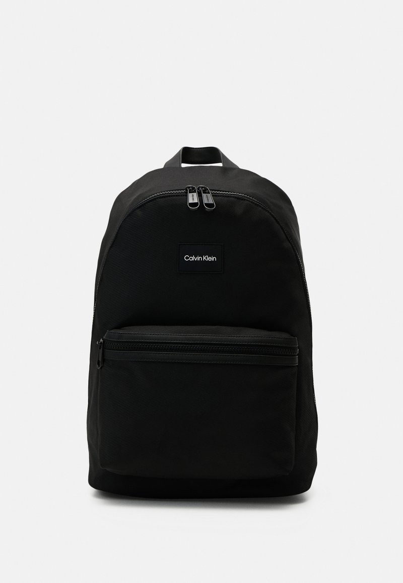 Zaino in tessuto nero con parte superiore arrotondata, doppie cerniere, tasca frontale, logo Calvin Klein e spallacci regolabili.