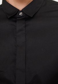 Gros plan d'un col de chemise noire boutonnée avec un seul bouton visible et un logo subtil "A|X" brodé sur la poitrine.