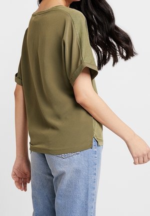 Woman with long dark hair wearing an olive green short-sleeve shirt and light blue jeans, shown from the back and side.