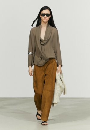 Frau geht, trägt Sonnenbrille, taupefarbene Bluse, braune weit geschnittene Hose, schwarze Sandalen, hält einen cremefarbenen Strickpullover in der Hand.