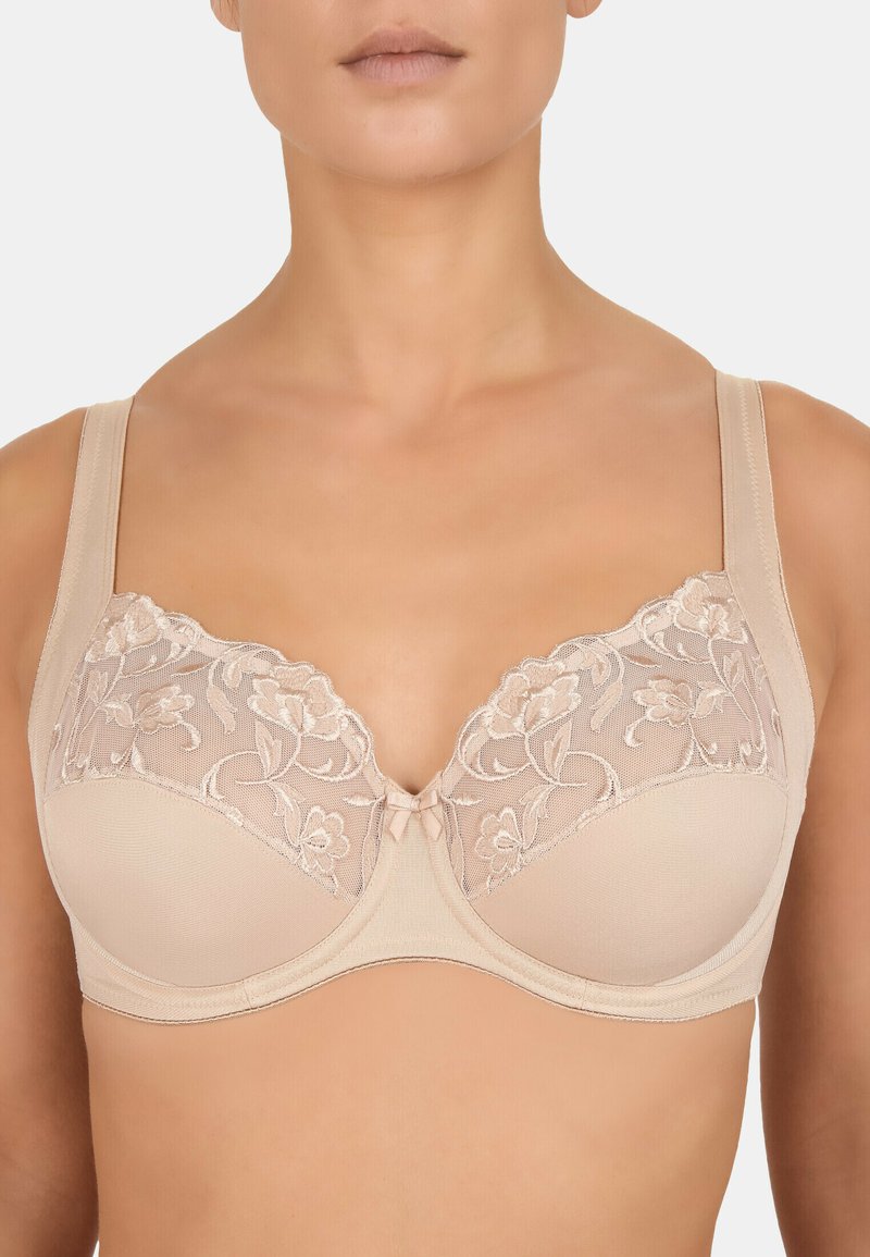 Soutien-gorge beige avec armatures, orné de dentelle florale, d'une bande lisse et de larges bretelles. Présente un petit nœud central comme embellissement.