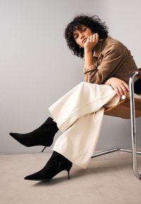 Black suede ankle boots with pointed toes and a thin heel, paired with cream wide-leg pants and a tan jacket, seated on a metal chair.