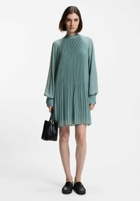 A pleated dress in muted green fabric with long sleeves and a high neckline, paired with a black handbag and black slip-on shoes.