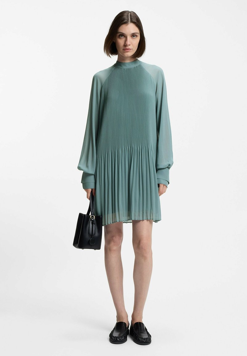 A pleated dress in muted green fabric with long sleeves and a high neckline, paired with a black handbag and black slip-on shoes.