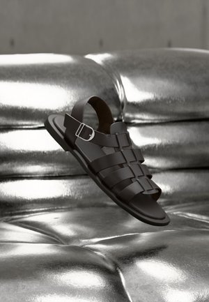 Schwarze Ledersandale für Herren mit verstellbarem Schnallenriemen, liegend auf einer glänzenden silbernen, gepolsterten Metalloberfläche.