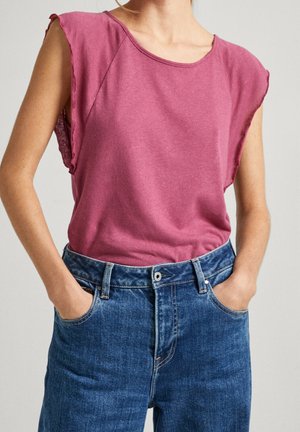 Femme portant un haut rose sans manches aux bords effilochés et un jean taille haute bleu, debout les mains dans les poches devant un fond uni.
