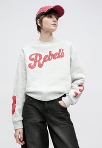 Pelēka džemperis ar uzrakstu "Rebels" treknā sarkanā fontā, ar garām piedurknēm un sarkanu ciparu 3 uz kreisās piedurknes, savienots ar tumšām džinsu biksēm.