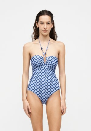 Femme aux longs cheveux foncés porte un maillot de bain une pièce bleu à motifs avec un lien noué autour du cou et une petite découpe en forme de trou à la poitrine.