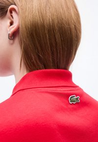 Άτομο με κοντά ανοιχτό καστανά μαλλιά, φορώντας κόκκινο πουκάμισο με ένα μικρό πράσινο λογότυπο κροκοδείλου κοντά στο γιακά στο πίσω μέρος.