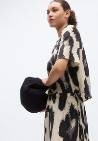 Čierny a krémový vzorovaný top s voľným strihom a krátkymi rukávmi, doplnený o zladený spodný diel. Drží čiernu taštičku. Textúrovaná látka.