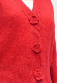 Sarkans kardigans no mīkstas, teksturētas vilnas ar V veida izgriezumu, ribotu apakšu un trim rožu formas pogām priekšpusē.