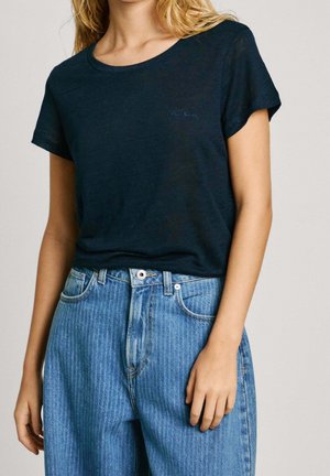 Žena má na sobě jednoduché tmavě modré tričko s krátkými rukávy zastrčené do světle modrých džínů s vysokým pasem a zapínáním na knoflíky.