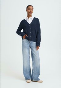 Marinblå stickad cardigan med V-ringning och stora framknappar, bärs över en vit skjorta och kombineras med ljusblå vida jeans.