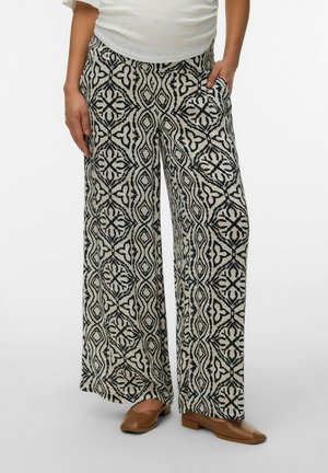 Sort-hvide bukser med vid ben og mønster, båret med en hvid top og brune flade sko med firkantet tå, den ene hånd i lommen.