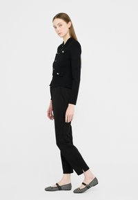Donna in giacca nera aderente con bottoni dorati, pantaloni neri slim e scarpe basse a quadretti bianchi e neri, in piedi di lato su uno sfondo semplice.