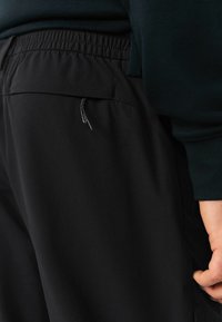 Nahaufnahme einer Person, die schwarze elastische Hose und ein schwarzes langärmliges Oberteil trägt, mit der rechten Hand entspannt an ihrer Seite.