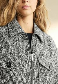 Veste grise et blanche texturée avec une fermeture éclair à l'avant, un grand col et deux boutons noirs sur les poches à rabat.
